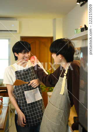 Cheerful gay couple wearing aprons preparing meal together in home kitchen Cheerful gay couple wearing aprons preparing meal together in home kitchen 122215009