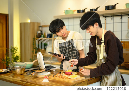 Cheerful gay couple wearing aprons preparing meal together in home kitchen 122215018