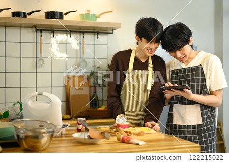 Cheerful gay couple wearing aprons preparing meal together in home kitchen 122215022