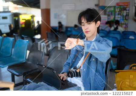 Young traveler checking time on his wristwatch while sitting at a bus station 122215027