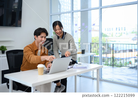 Two smiling coworkers reviewing project details on laptop at modern workplace 122215099