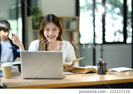 Cheerful young university student sitting at desk with laptop and open books Cheerful young university student sitting at desk with laptop and open books 122215138