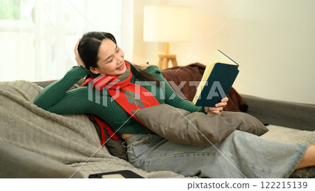 Cheerful young woman in green sweater and red scarf reading a book on couch Cheerful young woman in green sweater and red scarf reading a book on couch 122215139