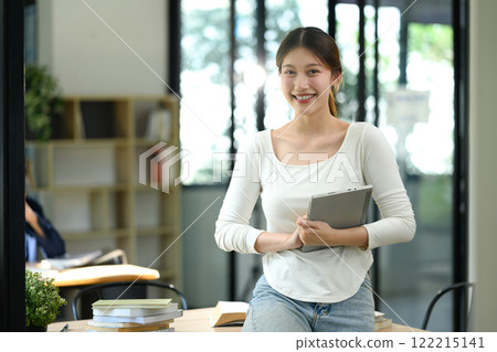 Portrait of cheerful university student holding digital tablet standing confidently in a modern study space Portrait of cheerful university student holding digital tablet standing confidently in a modern study space 122215141