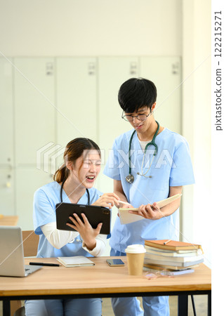 Two young medical students in blue scrubs reviewing a notes and using digital tablet during study session 122215271