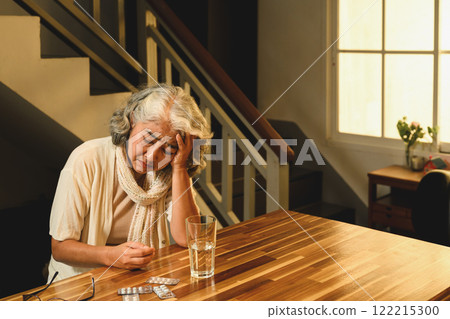 Mature woman holding head with a worried expression, sitting at a table with pills and a glass of water Mature woman holding head with a worried expression, sitting at a table with pills and a glass of water 122215300