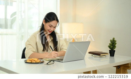 Smiling young woman writing in a notebook while working on her laptop 122215319