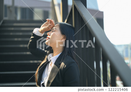 Stressed and exhausted businesswoman leaning against railing with hand on her forehead and closing her eyes Stressed and exhausted businesswoman leaning against railing with hand on her forehead and closing her eyes 122215406