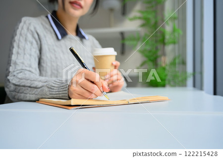 Close up of young woman writing in notebook while sitting in a bright, natural light filled space Close up of young woman writing in notebook while sitting in a bright, natural light filled space 122215428
