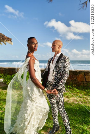 Ambiguous gender fluid black person in white dress, lgbtq groom in floral suit at tropical villa wedding. Tropical plants surrounds them at sunny beach ceremony celebration. 122216326
