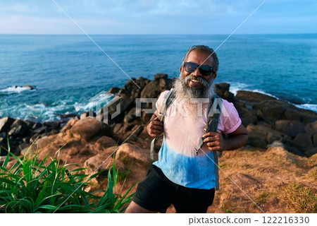 Senior man hikes by ocean cliffs. Older male admires sea view, smiles, holds backpack. Ethnic elder explores nature, enjoys outdoor adventure. Fit lifestyle, healthy aging in scenic landscape. Senior man hikes by ocean cliffs. Older male admires sea view, smiles, holds backpack. Ethnic elder explores nature, enjoys outdoor adventure. Fit lifestyle, healthy aging in scenic landscape. 122216330