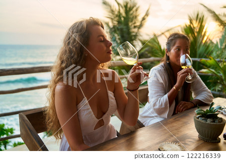 Two women enjoy wine in seaside restaurant. Friends relax, smile, sea view at sunset. Girls chat, taste drinks in outdoor setting. Afternoon leisure, conversation by ocean. 122216339