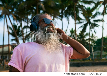 Smiling older man with headphones stands among palm trees. Enjoys tropical vibes, music in exotic setting. Casual wear, sunglasses. Relaxed outdoor mood. Sunlit beach, nature scene, coastal lifestyle. Smiling older man with headphones stands among palm trees. Enjoys tropical vibes, music in exotic setting. Casual wear, sunglasses. Relaxed outdoor mood. Sunlit beach, nature scene, coastal lifestyle. 122216340