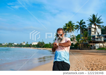 Senior ethnic man on beach uses smartwatch and wireless earbuds. Bearded old grandpa embraces modern tech innovations. Active lifestyle with wearable gadgets. Ocean backdrop creates calm vibe. Senior ethnic man on beach uses smartwatch and wireless earbuds. Bearded old grandpa embraces modern tech innovations. Active lifestyle with wearable gadgets. Ocean backdrop creates calm vibe. 122216348