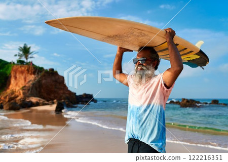 Senior man carries surfboard at beach. Active lifestyle for elderly. Grey-haired male enjoys ocean sport. Diverse mature surfer, vibrant life. Positive outlook, healthy aging. Senior man carries surfboard at beach. Active lifestyle for elderly. Grey-haired male enjoys ocean sport. Diverse mature surfer, vibrant life. Positive outlook, healthy aging. 122216351