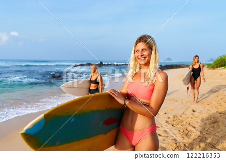 Three women on sandy beach holding surfboards smile, ready for surfing. Ocean waves in background. Summer vibe. Fitness and leisure focus. Surf camp scene. 122216353