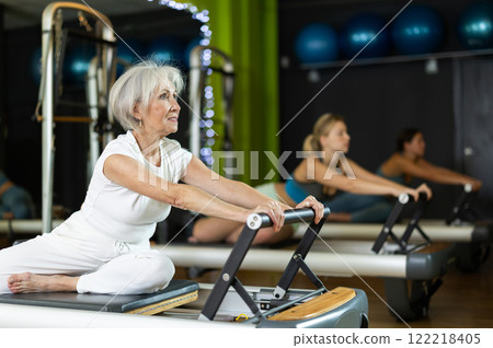 Mature woman sitting on foldable bed reformer performs exercises, strengthening body and muscles Mature woman sitting on foldable bed reformer performs exercises, strengthening body and muscles 122218405