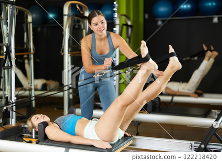 Girl trainer helps young woman to perform exercises in lying position on pilates reformer Girl trainer helps young woman to perform exercises in lying position on pilates reformer 122218420