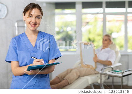 Young woman doctor posing in doctor's cabinet with patient lying on clinical chair 122218546