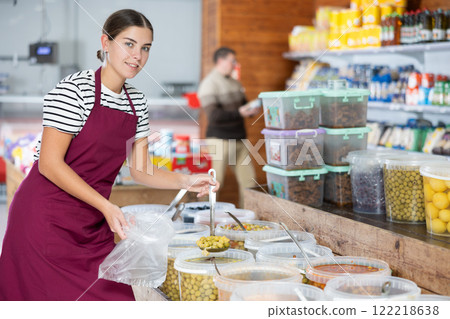 Friendly salesgirl filling cellophane bag with marinated olives for customer 122218638