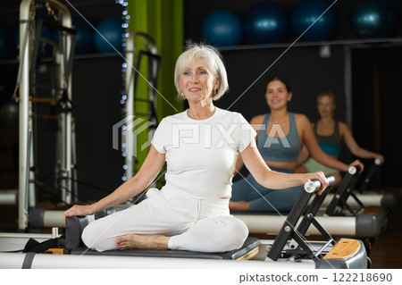 Mature woman sitting on foldable bed reformer performs exercises, strengthening body and muscles Mature woman sitting on foldable bed reformer performs exercises, strengthening body and muscles 122218690