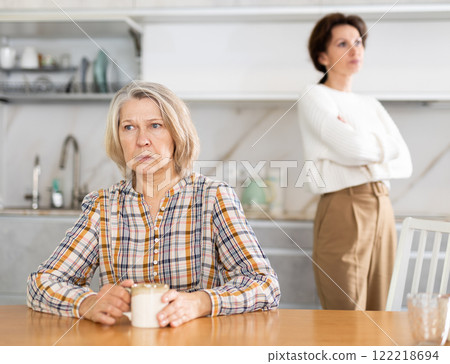 Upset old woman sitting at the kitchen table with her back to woman quarreling to her 122218694