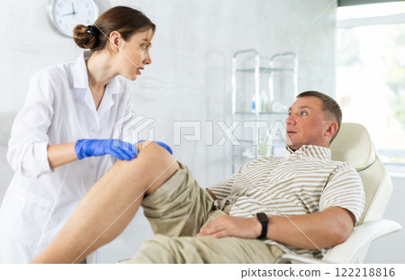 Man client of clinic looks at specialist and tells to woman doctor where knee hurts Man client of clinic looks at specialist and tells to woman doctor where knee hurts 122218816