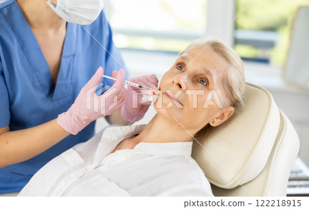 Professional cosmetologist performing lip filler injections to senior woman 122218915