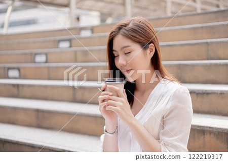 A young woman enjoys a warm beverage while sitting on outdoor steps. A young woman enjoys a warm beverage while sitting on outdoor steps. 122219317