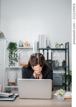 Businesswoman feeling stressed at her desk, working on a laptop in a modern office with plants and shelves. 122220928
