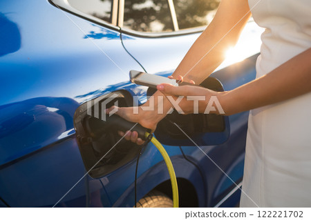 A woman connects an electric vehicle to a charger while checking her smartphone  122221702
