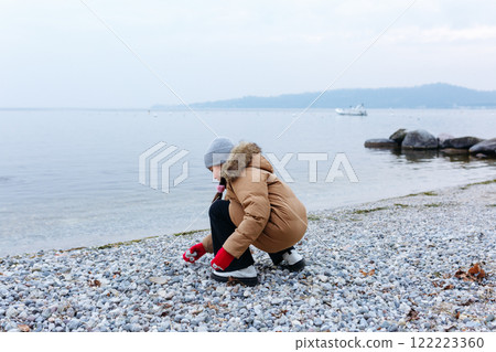 rear view of sitting on the beach girl in winter jacket playing with stones rear view of sitting on the beach girl in winter jacket playing with stones 122223360