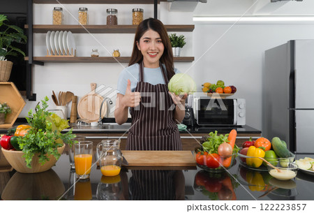 Young asian housewife dressed in an apron raise finger thumb up while holding fresh lettuce. Morning atmosphere in a modern kitchen. 122223857