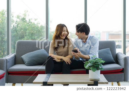 Young asian couple sitting on a couch, laughing together and sharing a mobile phone, in a bright living room. Young asian couple sitting on a couch, laughing together and sharing a mobile phone, in a bright living room. 122223942