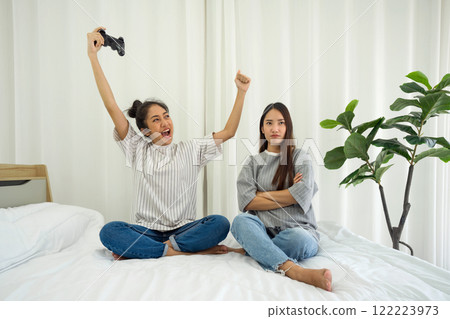 Two girl sit on a bed in a bright room. One girl raise her arm in excitement after winning a game, while the other appear annoyed, showcasing different reaction to gaming. Two girl sit on a bed in a bright room. One girl raise her arm in excitement after winning a game, while the other appear annoyed, showcasing different reaction to gaming. 122223973