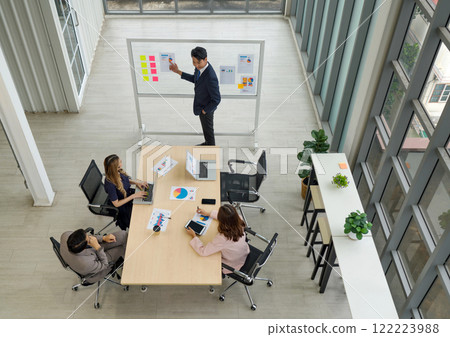 A business meeting in a modern office. A male presenter in suit pointing at colorful chart and graph as if he is delivering a report or presentation. 122223988