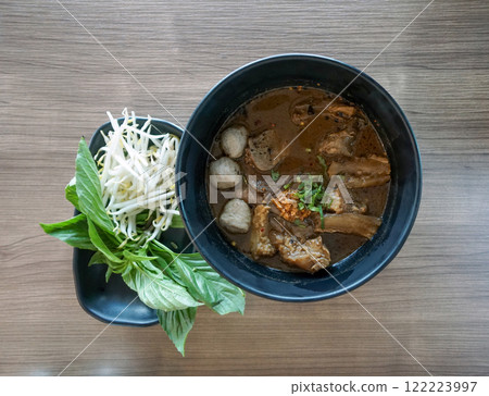 Premium thai style beef boat noodle in dark soy sauce, pickled bean curd, and some other spice soup served with meatball, tendon and braised shank. Fresh vegetable are on the table. Premium thai style beef boat noodle in dark soy sauce, pickled bean curd, and some other spice soup served with meatball, tendon and braised shank. Fresh vegetable are on the table. 122223997