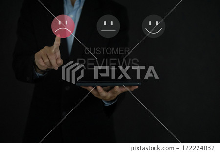 Businessman hand in black suit holding tablet computer, pointing at sad face icon to give dissatisfaction in service, rating very unimpressed. 122224032