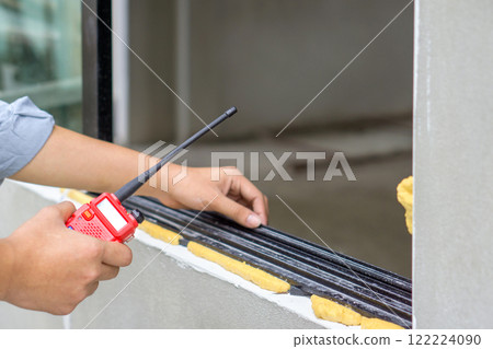 Closeup construction worker hand with walkie talkie near aluminium window frame. Work environment of engineer at the construction site of housing projects. 122224090