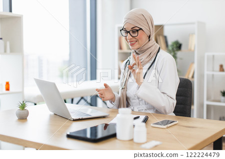 Young Muslim female doctor having video call with patient using laptop in modern office. Middle Eastern woman illustrating healthcare communication technology. Young Muslim female doctor having video call with patient using laptop in modern office. Middle Eastern woman illustrating healthcare communication technology. 122224479