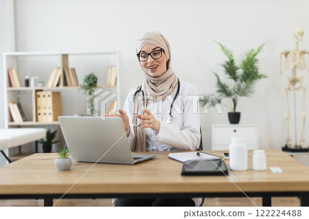 Middle-Eastern female doctor wearing hijab conducting online consultation using laptop. Focus on telemedicine, remote healthcare. Modern and professional office setting enhances environment. Middle-Eastern female doctor wearing hijab conducting online consultation using laptop. Focus on telemedicine, remote healthcare. Modern and professional office setting enhances environment. 122224488