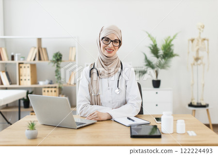 Smiling young Middle Eastern female doctor in hijab sitting at desk in modern medical office with laptop and notepad showcasing professionalism 122224493