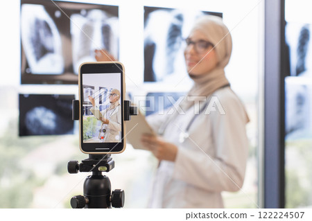 Muslim woman doctor records video lesson using smartphone and x-rays. Middle Eastern female professional teaches healthcare concepts. Online education setting with focus on technology and knowledge. Muslim woman doctor records video lesson using smartphone and x-rays. Middle Eastern female professional teaches healthcare concepts. Online education setting with focus on technology and knowledge. 122224507