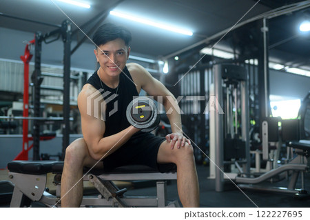 Strong handsome sportsman man sitting on a gym bench and lifting a dumbbell 122227695