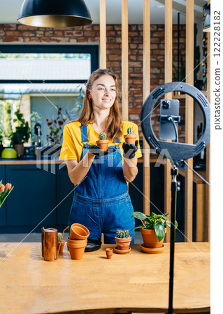 Positive woman holing potted plants and demonstrates her results with videocall on smartphone transplanting enjoys her hobby shares images in social network wears yellow t-shirt, overalls at home. Positive woman holing potted plants and demonstrates her results with videocall on smartphone transplanting enjoys her hobby shares images in social network wears yellow t-shirt, overalls at home. 122228182