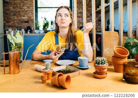 Searching for best advice. Romantic brunette haired smiling female gardener eats sweet cake, drinking coffee sitting surround her plants pots, equipment, tools watering can in home garden at home. Searching for best advice. Romantic brunette haired smiling female gardener eats sweet cake, drinking coffee sitting surround her plants pots, equipment, tools watering can in home garden at home. 122228200