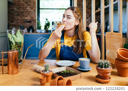 Searching for best advice. Romantic brunette haired smiling female gardener eats sweet cake, drinking coffee sitting surround her plants pots, equipment, tools, watering can in home garden at home. Searching for best advice. Romantic brunette haired smiling female gardener eats sweet cake, drinking coffee sitting surround her plants pots, equipment, tools, watering can in home garden at home. 122228201