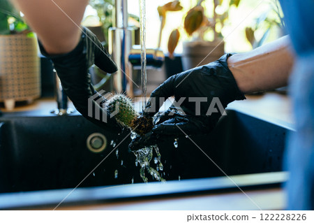 Close up of unrecognizable woman gardener wearing black gloves washing rinses the cactus root from the soil in kitchen. Gardening at home concept. Close up of unrecognizable woman gardener wearing black gloves washing rinses the cactus root from the soil in kitchen. Gardening at home concept. 122228226
