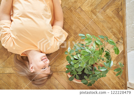 Caucasian female young lying on wooden floor smiling beside green potted plant 122228479