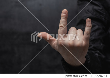 Close-up of a man in a shirt shows hand gesture Rock'n'roll against a concrete wall. Close-up of a man in a shirt shows hand gesture Rock'n'roll against a concrete wall. 122228781